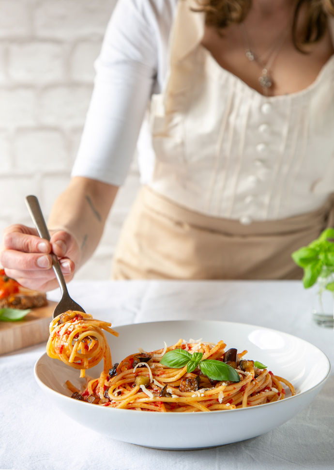 cook holding a fork with pasta over a bowl of pasta alla norma topped with basil leaves