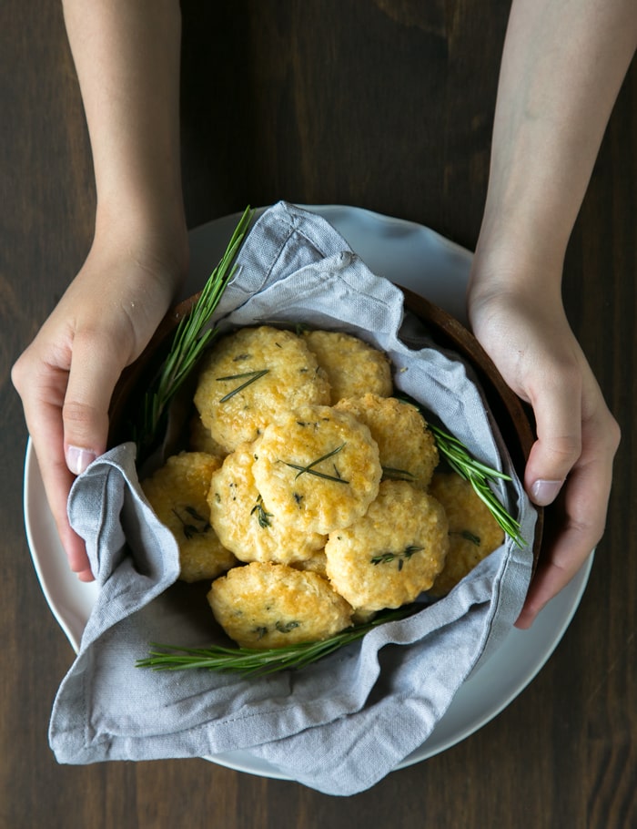 hands holding a wood bowl covered with napkin and filled with parmesan cookies and rosemary sprigs, wood board in the background