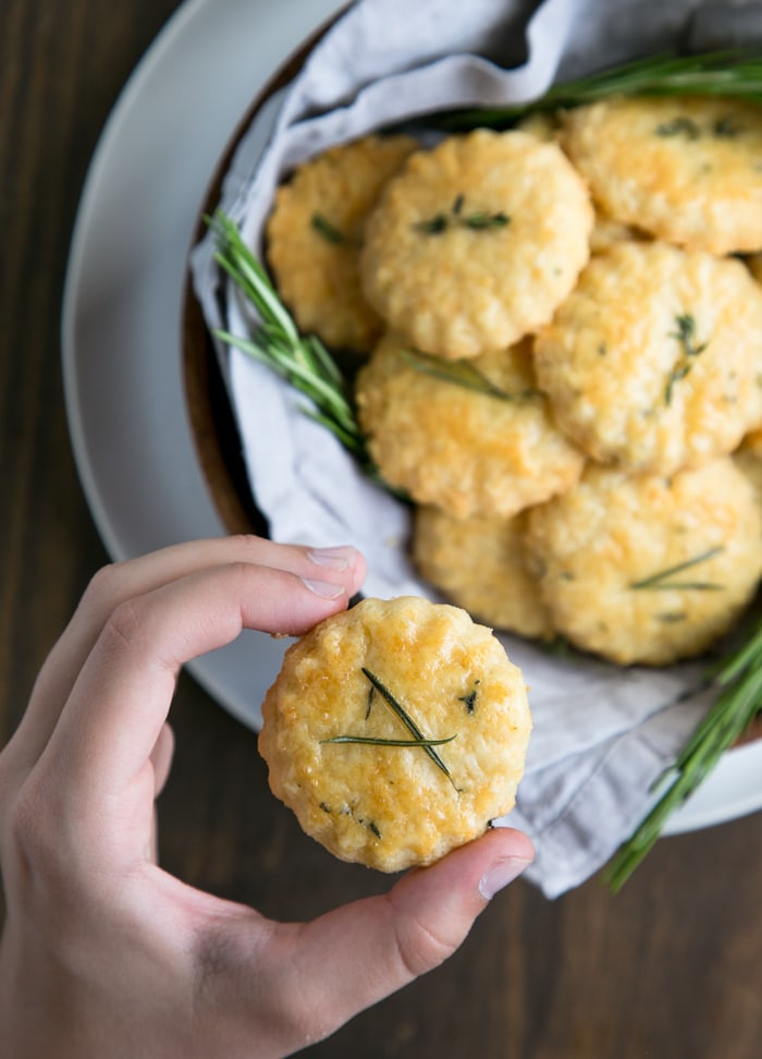 hand holding a parmesan cookies, plate with parmesan cookies and rosemary sprigs in the background.