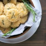 parmesan cookies and rosemary sprigs in a wood bowl covered with grey napkin, white plate beneath on wood table.