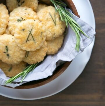 parmesan cookies and rosemary sprigs in a wood bowl covered with grey napkin, white plate beneath on wood table.