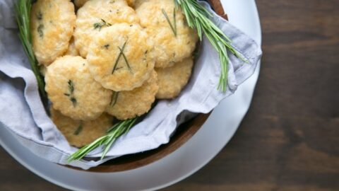 parmesan cookies and rosemary sprigs in a wood bowl covered with grey napkin, white plate beneath on wood table.