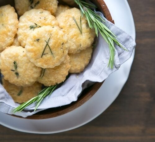 parmesan cookies and rosemary sprigs in a wood bowl covered with grey napkin, white plate beneath on wood table.