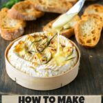 image optimized for pinterest: baked camembert in wooden box on a wooden board, knife dripping melted cheese, toasted baguette slices and sprigs of rosemary in the background