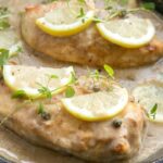 Chicken piccata in a large skillet with sauce, topped with lemon slices and thyme leaves. Image for pinterest