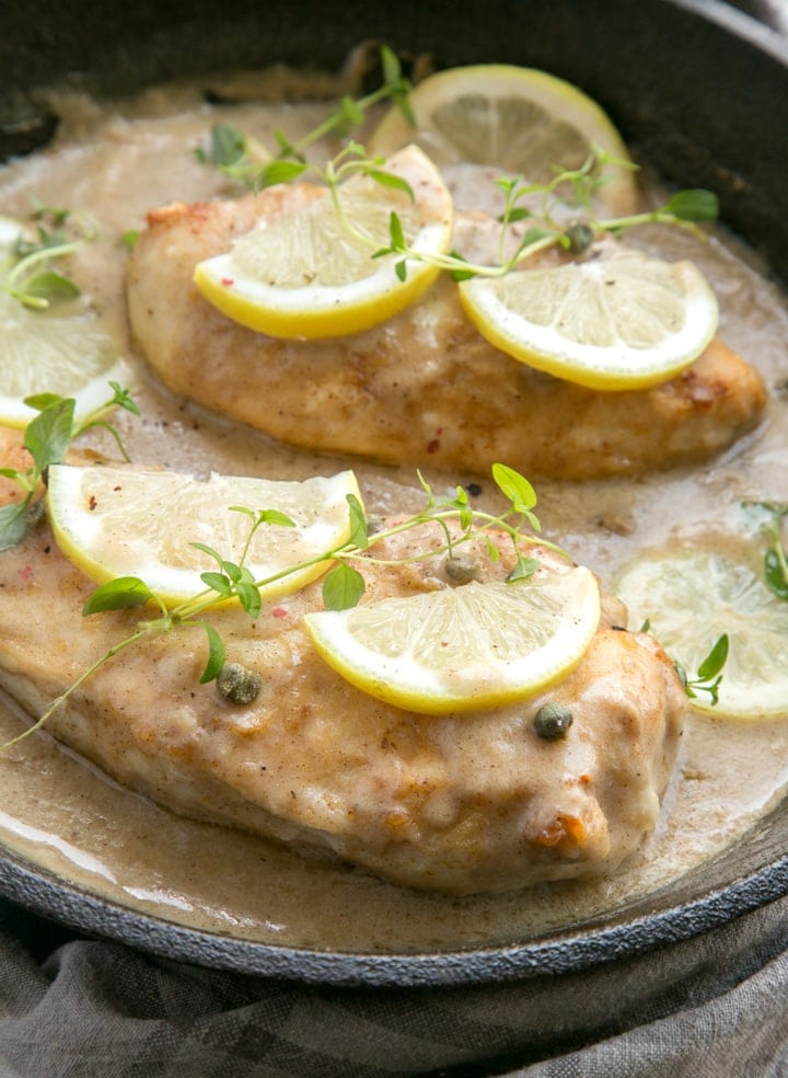 Chicken piccata in a large skillet with sauce, topped with lemon slices and thyme leaves