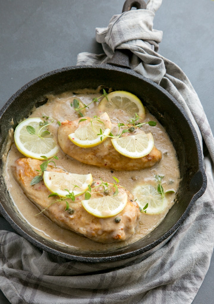 Chicken piccata in a large cast iron skillet with sauce, topped with lemon slices and thyme leaves, grey napkin underneath the skillet