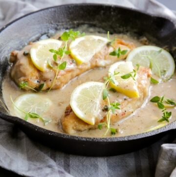 Chicken piccata in a large skillet with sauce, topped with lemon slices and thyme leaves