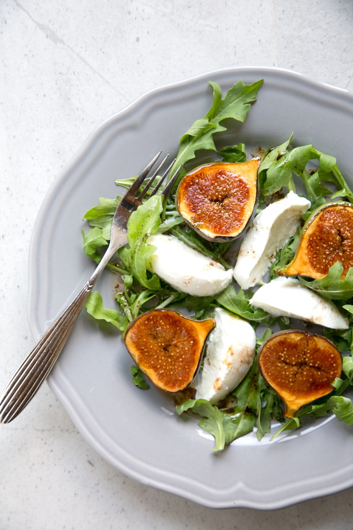 fig and mozzarella salad with arugula on grey plate with fork on th left and knife on the right, two fresh figs on the side over grey napkin