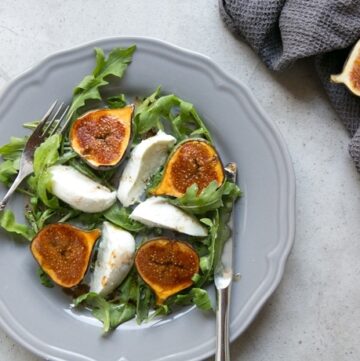 fig and mozzarella salad with arugula on grey plate with fork on th left and knife on the right, two fresh figs on the side over grey napkin