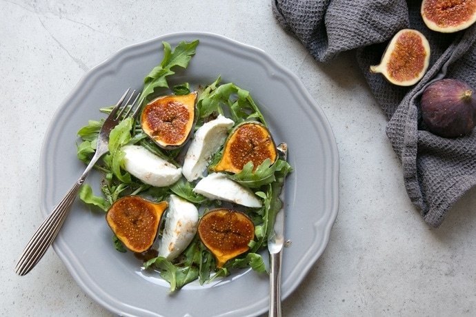fig and mozzarella salad with arugula on grey plate with fork on th left and knife on the right, two fresh figs on the side over grey napkin