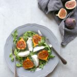 fig and mozzarella salad with arugula on grey plate with fork on th left and knife on the right, two fresh figs on the side over grey napkin