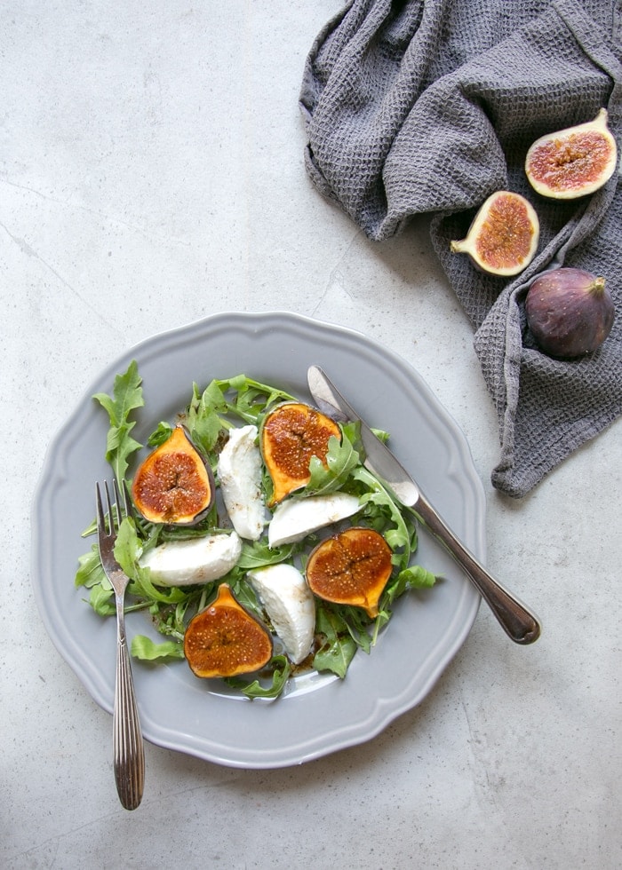 fig and mozzarella salad with arugula on grey plate with fork on th left and knife on the right, two fresh figs on the side over grey napkin