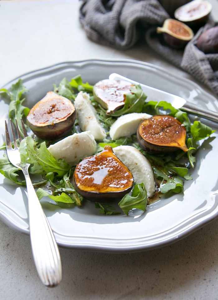 fig and mozzarella salad with arugula on grey plate with fork on th left and knife on the right, two fresh figs on the side over grey napkin