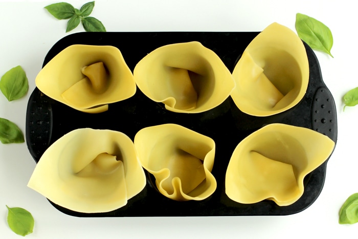  cooked lasagna sheet fitted in a muffin tin, basil leaves scattered all around in the white background