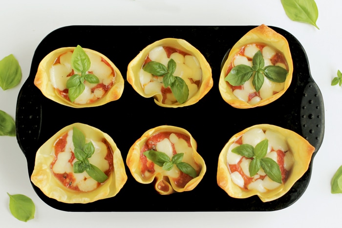 lasagna cups fitted in a muffin tin, basil leaves scattered all around in the white background