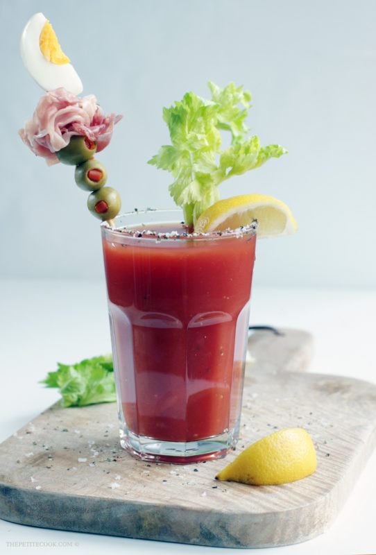 Bloody mary cocktail, topped with lemon wedge, and a skewer with three olives, one prosciutto slice, a wedge of boiled egg.