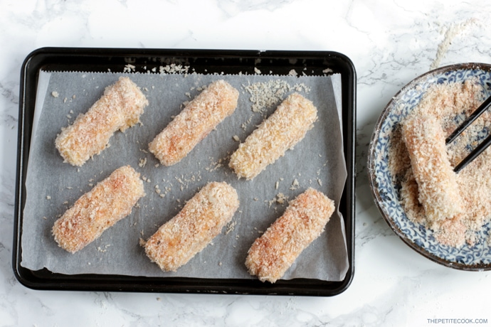 recipe process step 3: mozzarella sticks dipped in flour, eggs and panko breadcrumbs then arranged on a baking tray covered with parchment paper