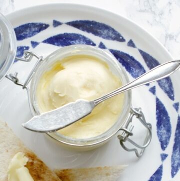 homemade butter in jar with knife over it, bread toasted with butter on top on a large white and blue plate, image optimized for Pinterest