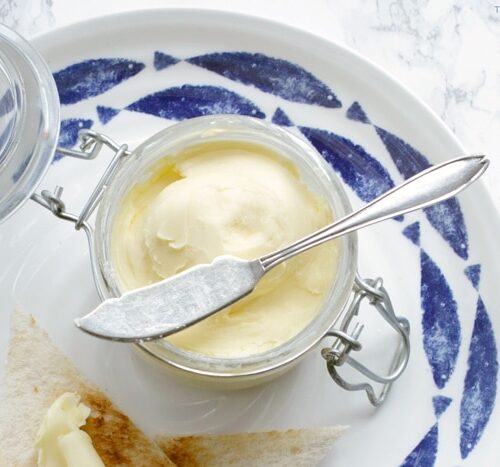 homemade butter in jar with knife over it, bread toasted with butter on top on a large white and blue plate, image optimized for Pinterest