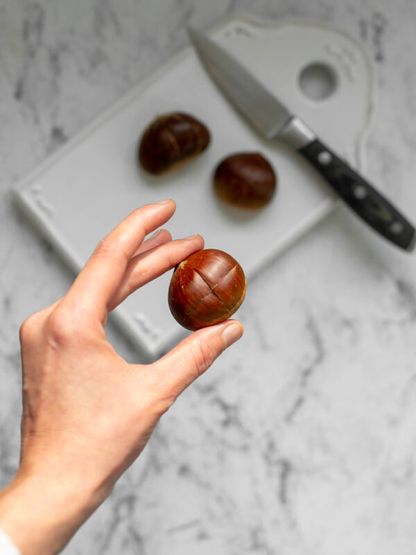 hand holding a chestnut scored with a cross. in the background a chopping board with a pairing knife and 2 chestnuts on top.