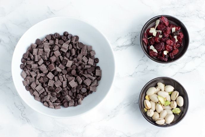 chocolate chips in one bowl, next to dried cranberries in a small bowl, next to pistachios into another small bowl.