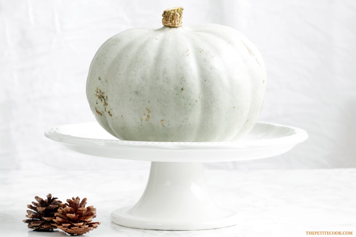 white pumpkin on a white cake stand next to two pine cones.