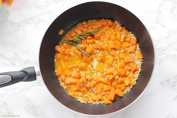 recipe step 3: barley, stock, pumpkin, garlic and rosemary sprigs simmering in a skillet.