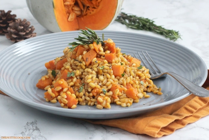Pumpkin pearl barley risotto on a grey plate topped rosemary sprig, and an open cut pumpkin in the background.