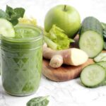 morning green juice in a glass, next to a wood board with fresh ginger, celery stalks, cucumber and cucumber slices, green apple and spinach leaves