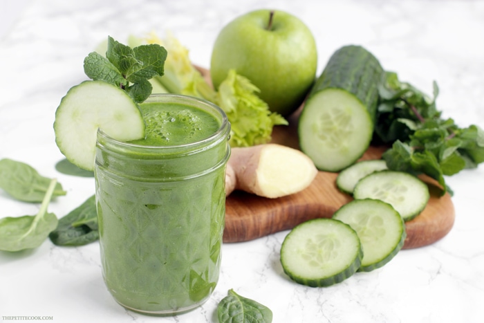 green juice in a glass, next to a wood board with fresh ginger, celery stalks, cucumber and cucumber slices, green apple and spinach leaves