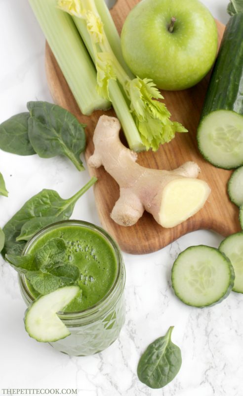 homemade juice in a glass, next to a wood board with fresh ginger, celery stalks, cucumber and cucumber slices, green apple and spinach leaves