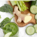 Image optimized for pinterest: morning green juice in a glass, next to a wood board with fresh ginger, celery stalks, cucumber and cucumber slices, green apple and spinach leaves