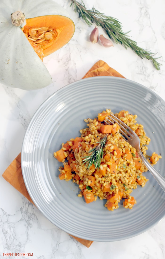 Barley Risotto with Pumpkin and Rosemary in a grey plate over orange napkin, cut pumpkin garlic cloves and rosemary sprigs in the background.