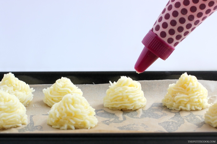 recipe step 2: unbaked duchess potatoes on a baking tray covered with parchment paper, next a piping bag