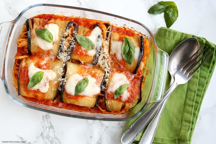 Eggplant spaghetti sandwiches in baking dish next to green napkin with spoon and fork on top, basil leaves on the side