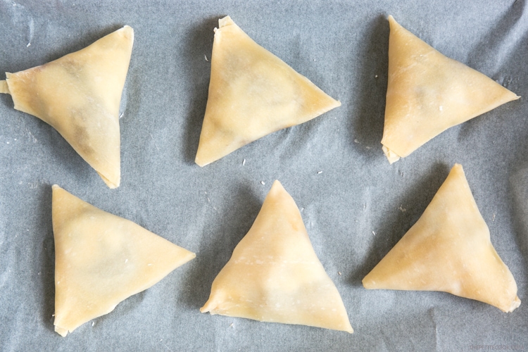 recipe step 3: samosas on a baking sheet covered with parchment paper, ready to be baked in the oven.