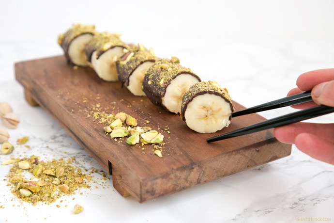 Pistachio Chocolate Banana Sushi on a wood board, hand holding chopstick on the right side.