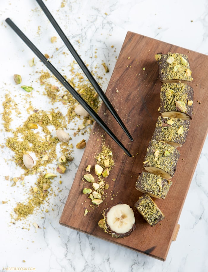 chocolate banana sushi with pistachios on top on a wood board, with chopsticks on the side, and crumbled pistachio in the background