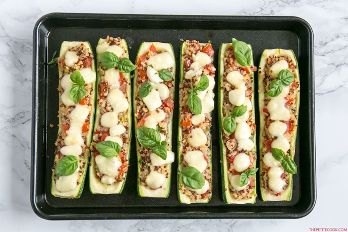 zucchini boats stuffed with quinoa and pico de gallo, topped with melted mozzarella cheese and basil leaves, arranged on a baking tray