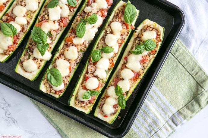 Zucchini boats stuffed qith quinoa and pico de gallo, topped with melted mozzarella cheese and basil leaves, arranged on a baking tray with green napkin beneath