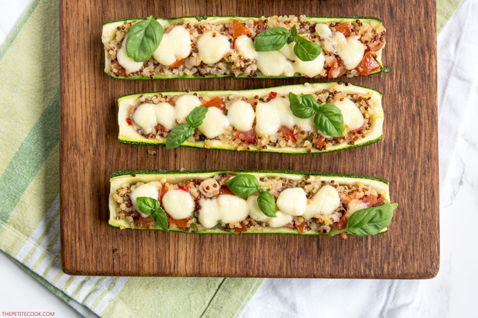 Zucchini boats stuffed qith quinoa and pico de gallo, topped with melted mozzarella cheese and basil leaves, on wood board with green napkin beneath