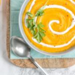 spicy pumpkin and carrot soup in a bowl, with light blue napkin and wood board beneath, small pumpkin on the left side