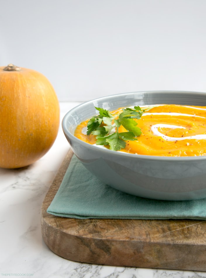 vegan pumpkin and carrot soup in a bowl, with light blue napkin and wood board beneath, small pumpkin on the left side 