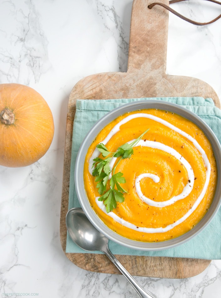 Vegan Pumpkin and Carrot soup topped with yogurt and coriander stem, in a grey plate over light blue napkin and wood board, next to a spoon and pumpkin