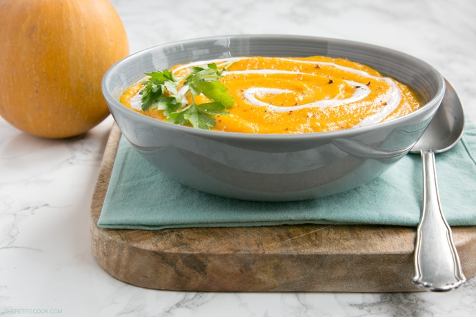 vegan pumpkin and carrot soup in a bowl, with light blue napkin and wood board beneath, small pumpkin on the left side 