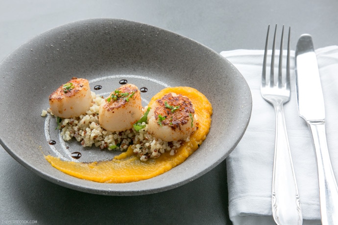 spicy scallops with pumpkin puree in a grey plate with fork and knife on the side on a white napkin