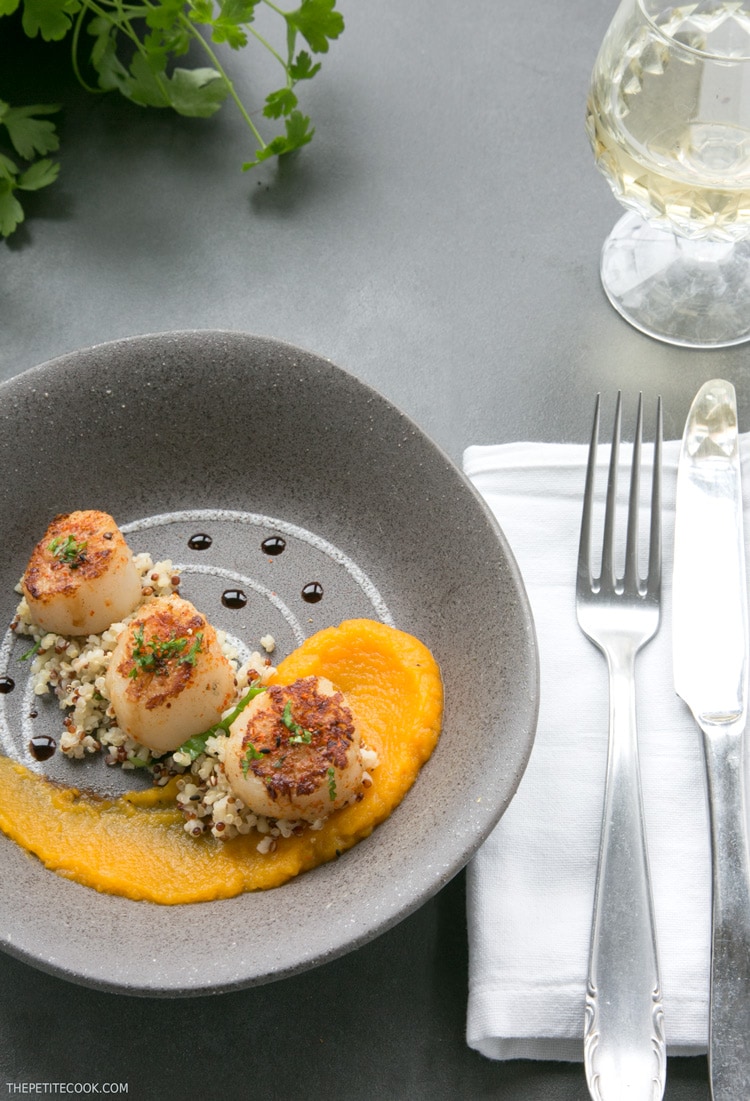 spicy scallops with pumpkin puree in a grey plate with fork and knife on the side on a white napkin and white wine glass on top right