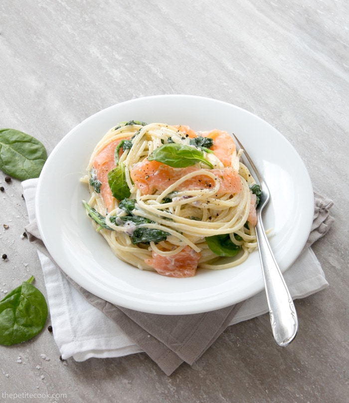 Spaghetti with Salmon and Spinach in a white plate with a fork, over a grey and a white napkins, spinach leaves pepper and salt scattered next to the plate