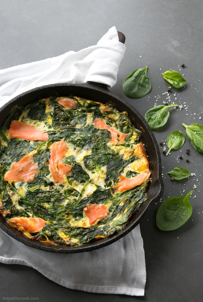 Crustless quiche with salmon and spinach in a cast iron skillet over a white napkin, next to the skillet spinach leaves, salt and pepper scattered over